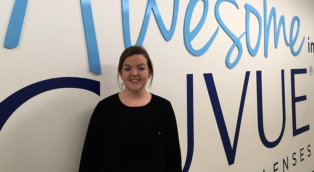 A young woman with brown hair tied back stands smiling in front of a large Acuvue sign in Johnson & Johnson Vision Care.