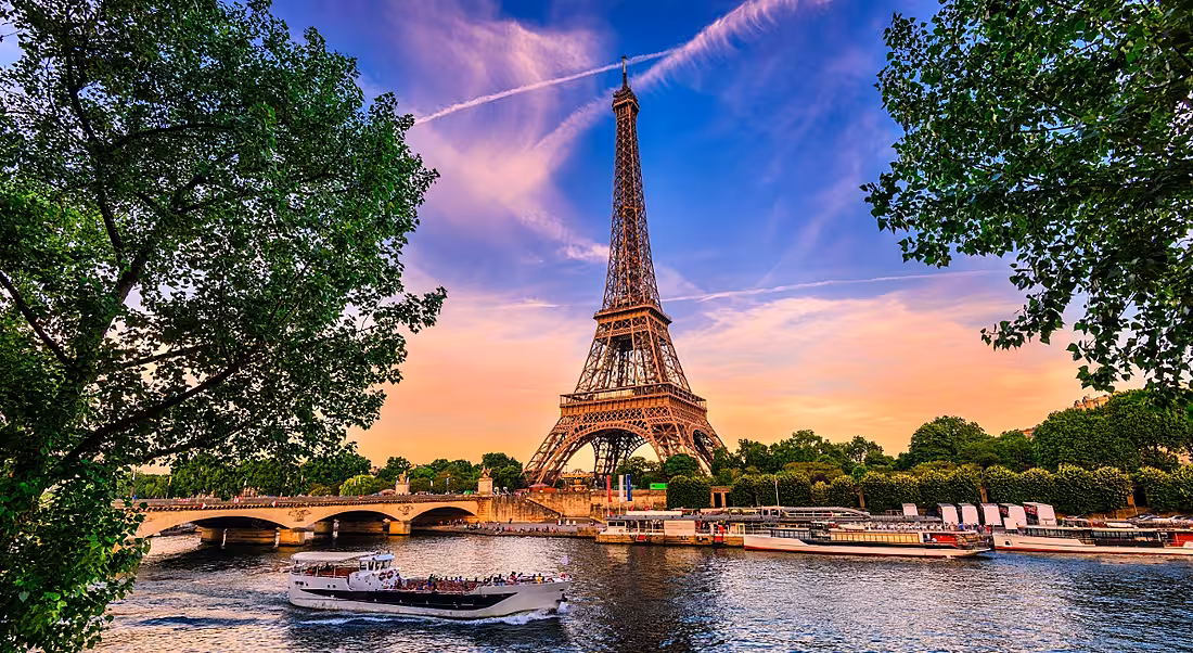 A view of golden Eiffel Tower against purple sunset backdrop, with green trees and canal boats in foreground.