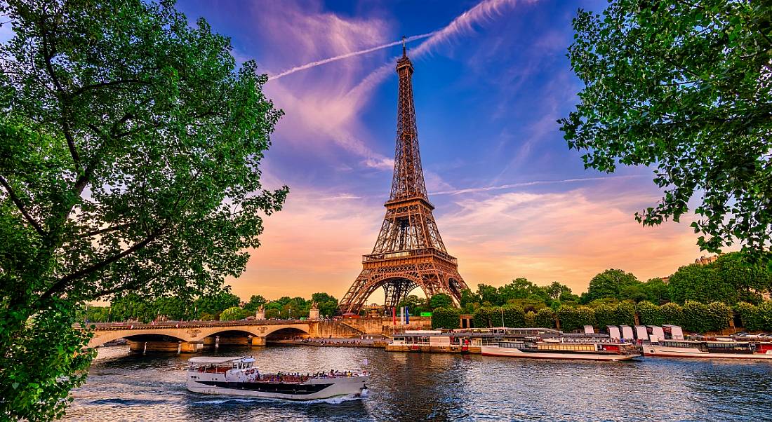 A view of golden Eiffel Tower against purple sunset backdrop, with green trees and canal boats in foreground.