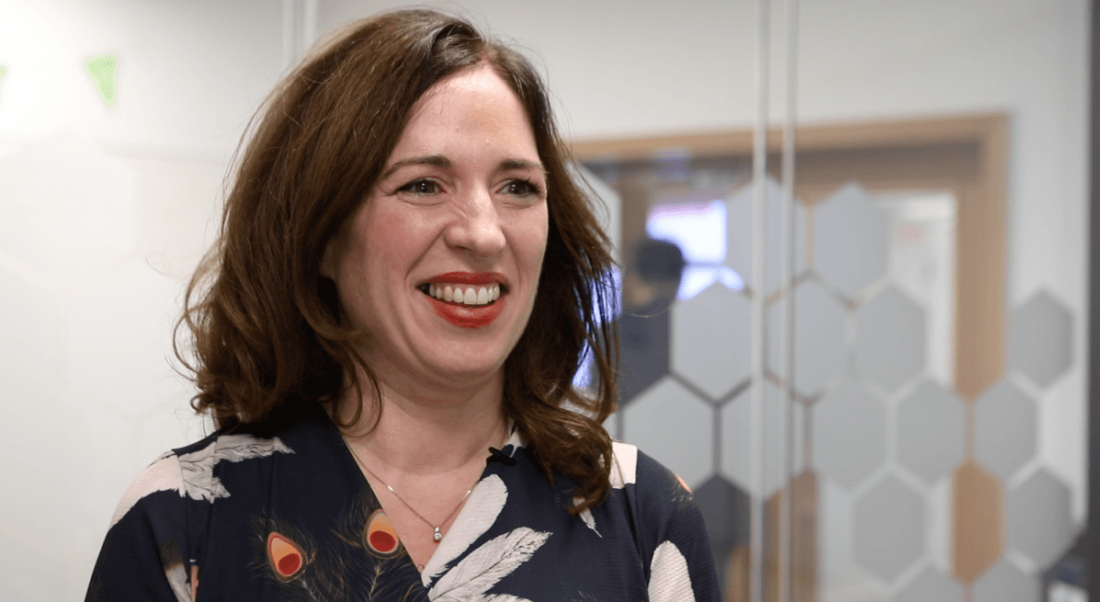 A woman with brown hair wearing a navy, flowery dress and smiling in the ACIA offices.