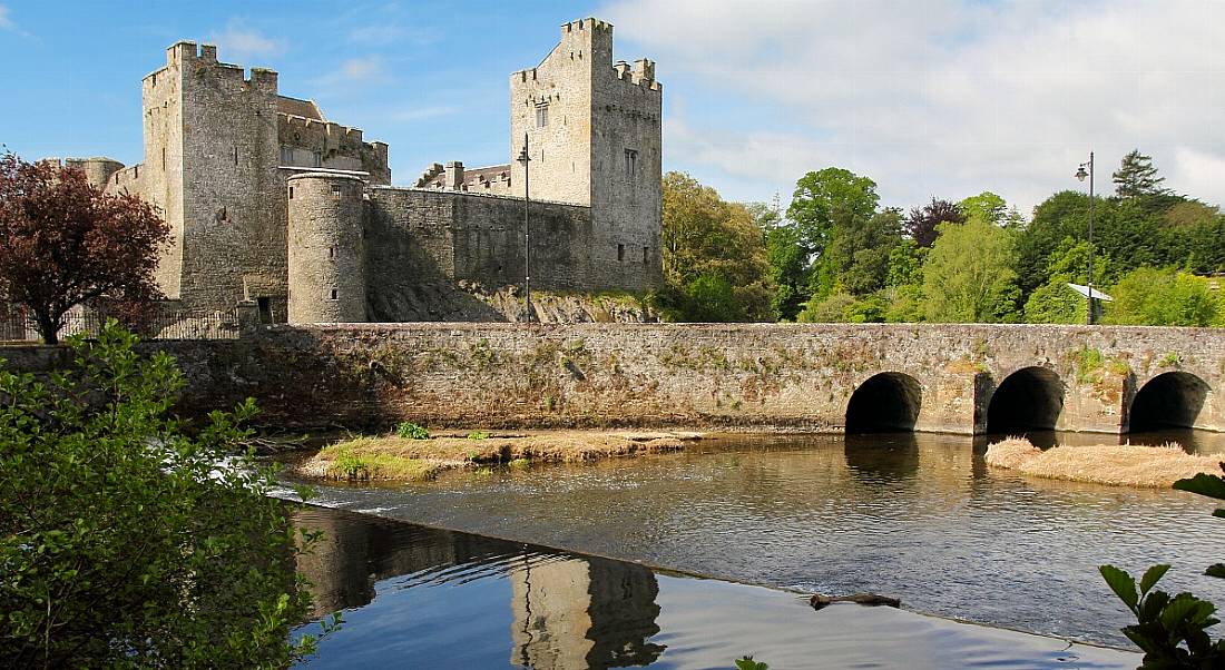 Grey stone castle of Cahir in Tipperary reflected in water under bridge in foreground.