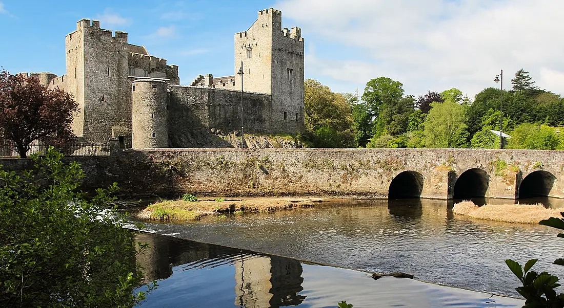 Grey stone castle of Cahir in Tipperary reflected in water under bridge in foreground.