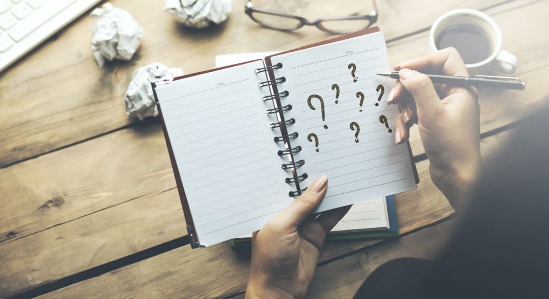 A notebook someone is holding. They are writing question marks on it representing questions after a job interview.