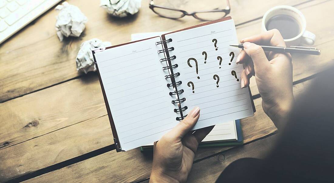 A notebook someone is holding. They are writing question marks on it representing questions after a job interview.