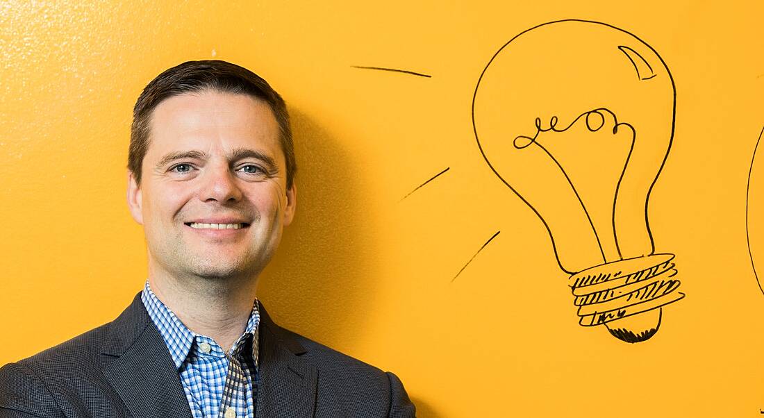 A man in a grey suit and checked shirt smiling at the camera against a canary yellow wall with a lightbulb motif.