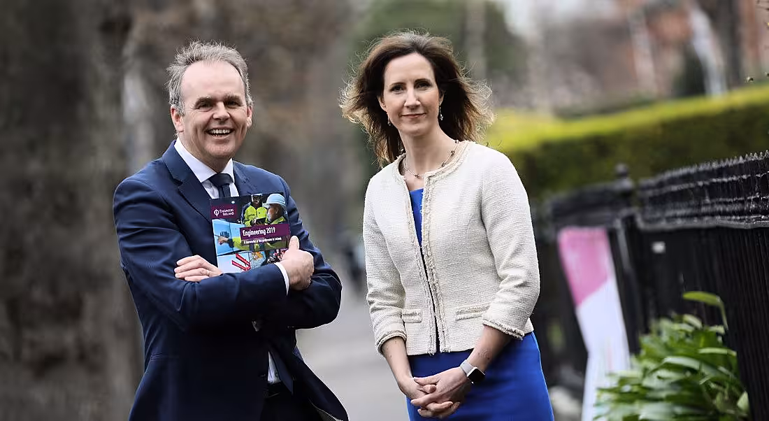 A man in a suit holds the Engineering 2019 report with his arms folded, next to a smartly dressed woman. They pose for a photo outside beside a railing.