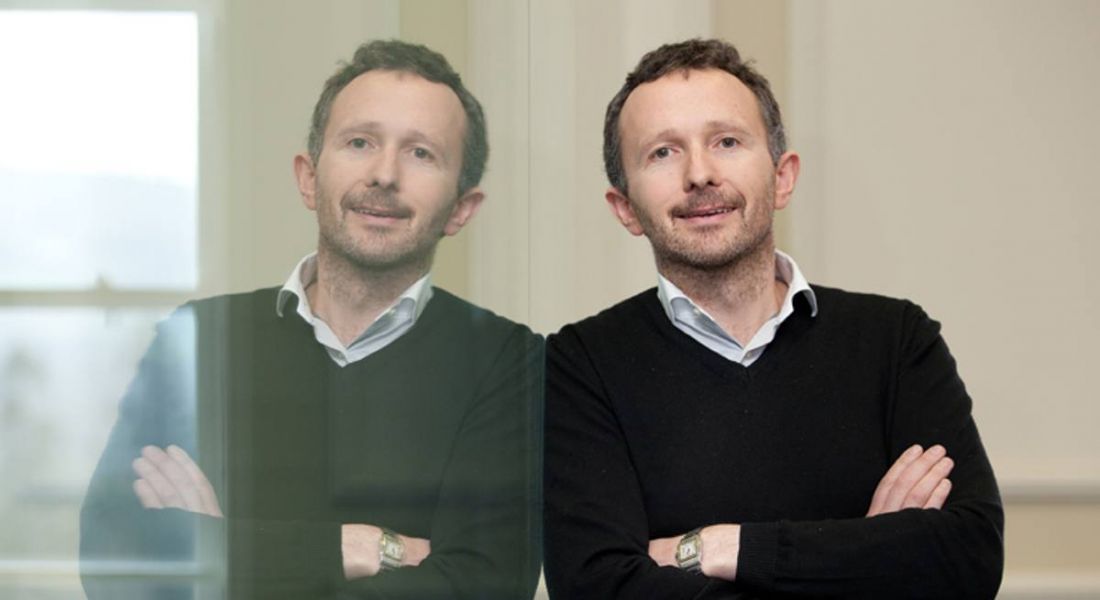 A man in a black sweater and white shirt leaning against a glass wall that is mirror reflecting his image.