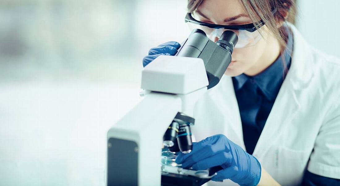 A female scientist wearing lab gear looking through a microscope and doing experiments.