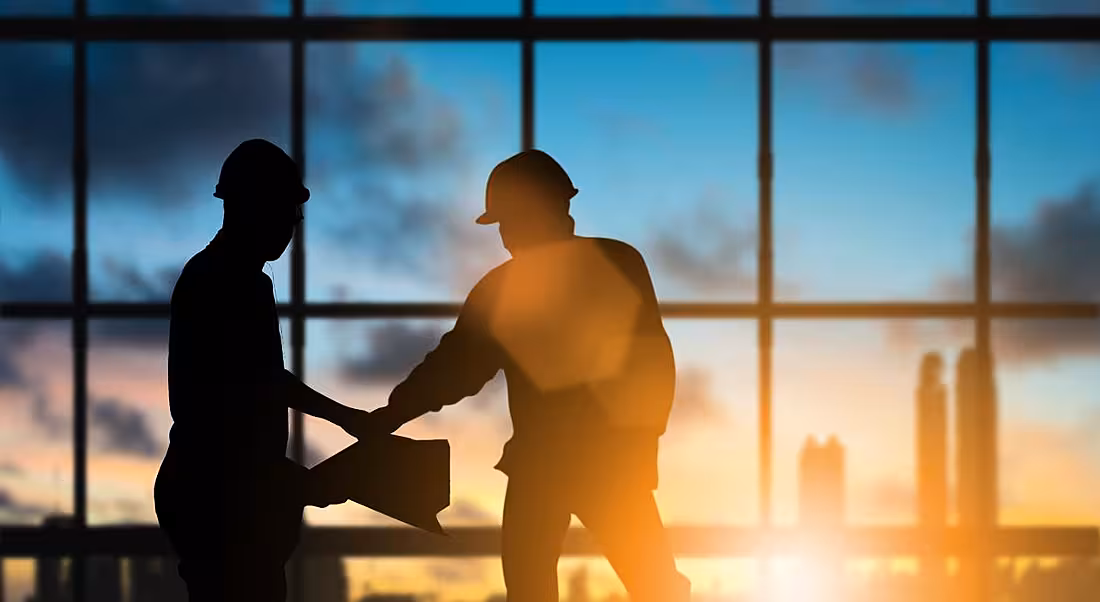 Silhouette of engineers in hard hats looking at building plans against the backdrop of a floor-to-ceiling window.