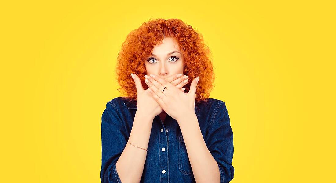 Against a bright yellow background, a woman with curly red hair wearing a denim shirt covers her mouth with her hands.