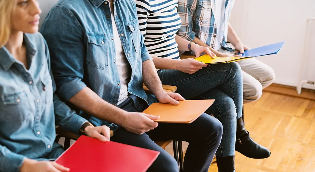 A group of people holding CVs in folders sitting in line against a wall waiting to be called for interview.