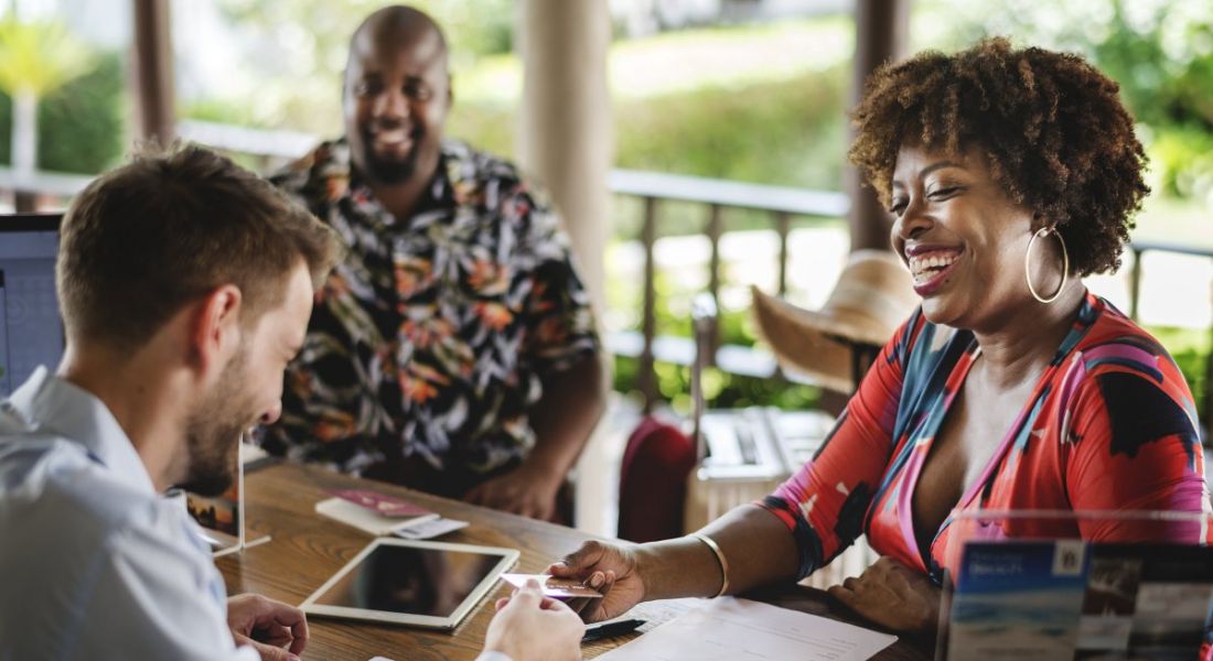 A group of happy employees laughing together sitting around a dark wood table on a patio with neocolonial columns in the background.