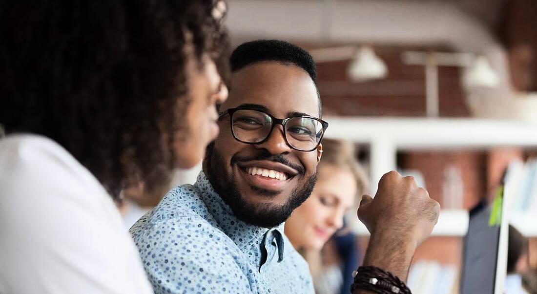 View of a young smiling man wearing thick-rimmed glasses looking at his co-worker.