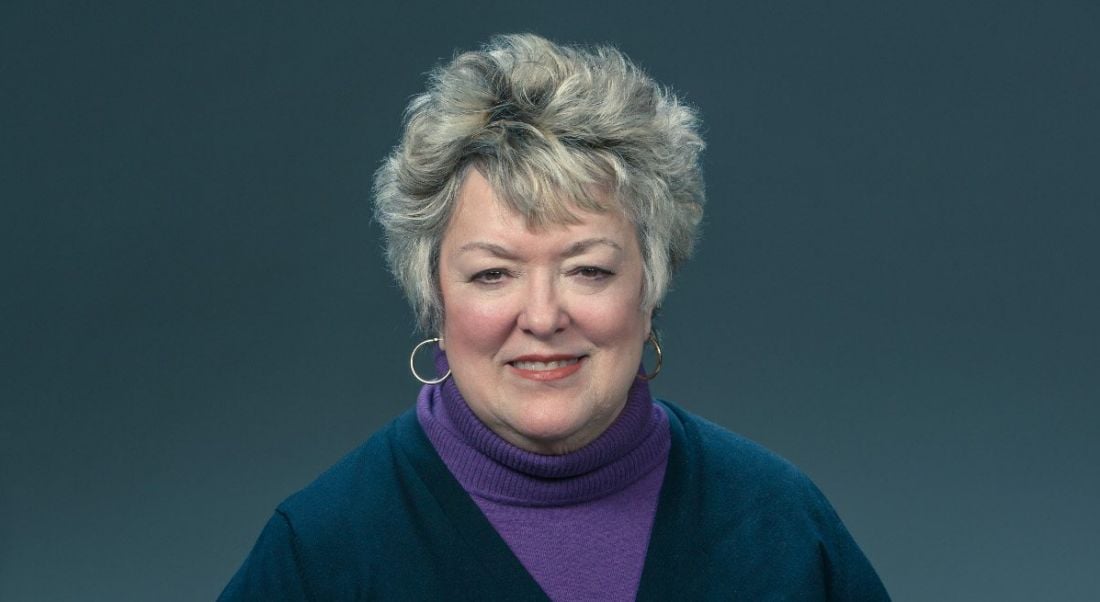 A close-up of a smiling woman with short grey hair wearing a navy cardigan and purple turtleneck. She has had a long cybersecurity career.