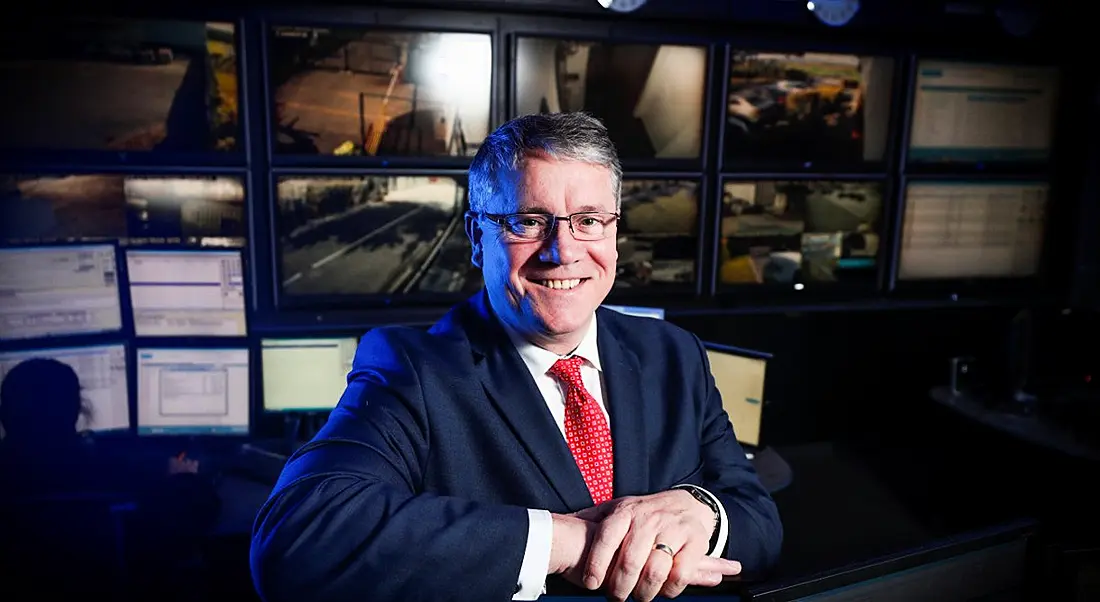 Man with dark suit, greying hair and glasses, sitting in a security control room.