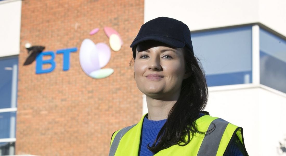 A woman with long dark hair wearing hi-vis vest and dark baseball cap stands outside a BT building.