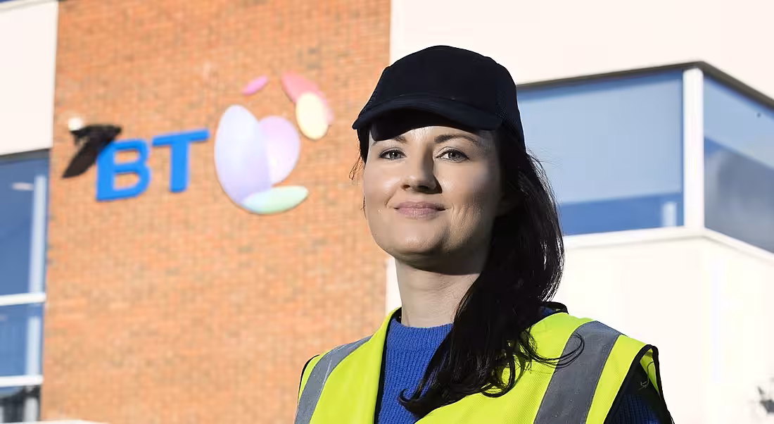 A woman with long dark hair wearing hi-vis vest and dark baseball cap stands outside a BT building.