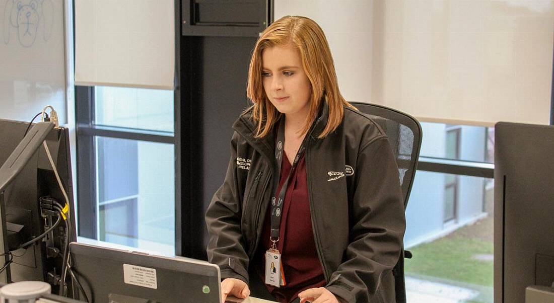 A red-haired young woman in a Jaguar Land Rover jacket working at a laptop.