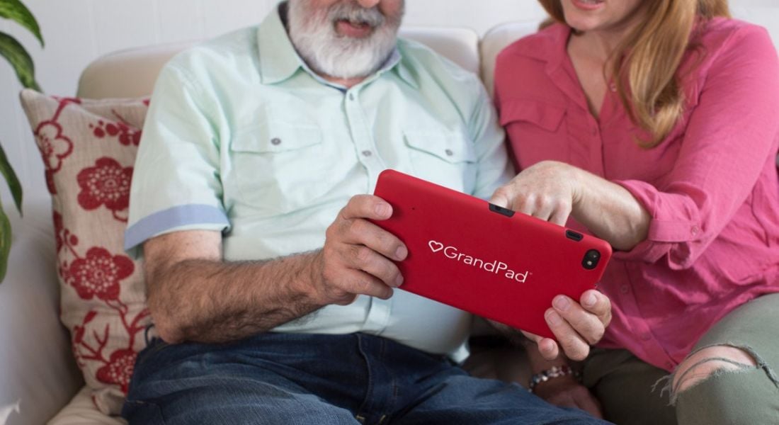 Elderly man being shown how to use a red tablet computer by a younger woman.