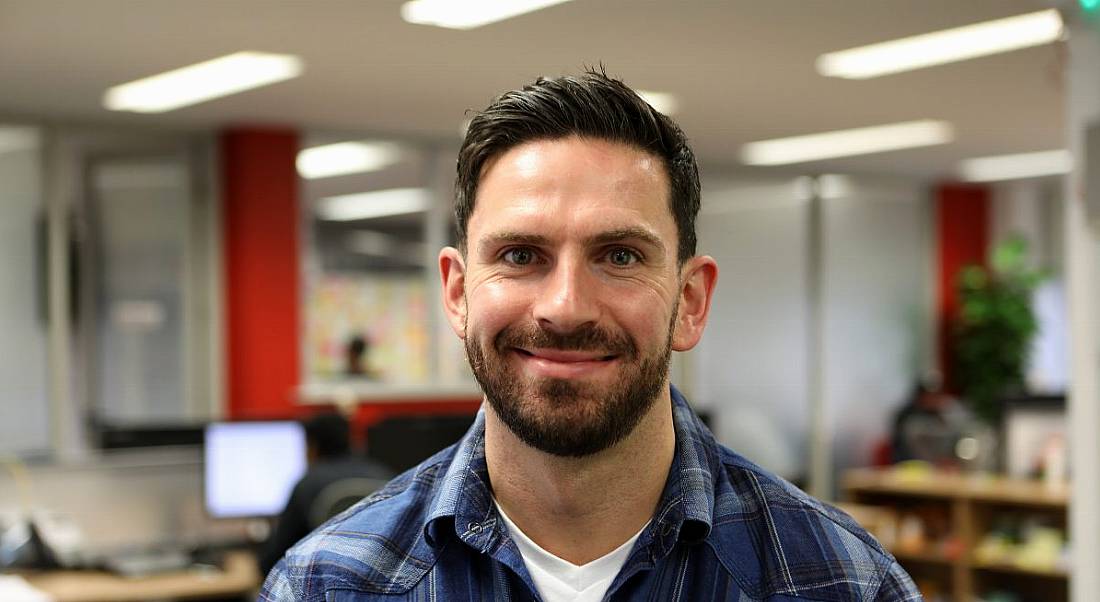 A young dark-haired man in a flannel shirt looks directly at the camera and smiles widely, showing dimples.