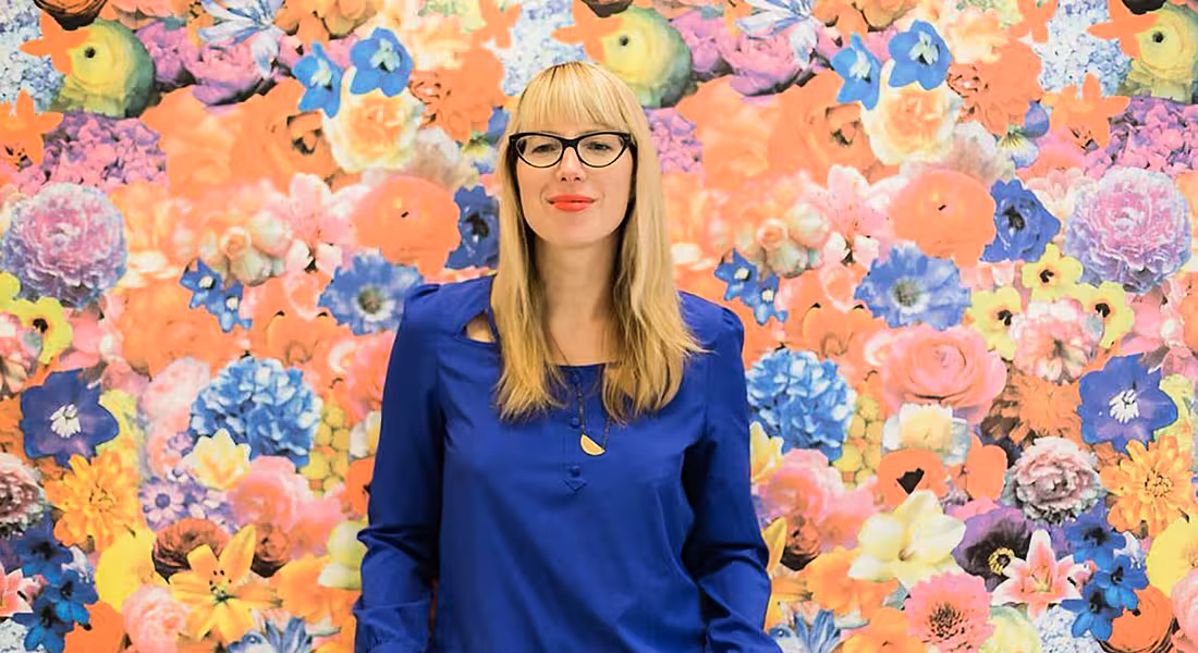 A blonde woman with glasses wearing a blue top standing in front of a floral backdrop. She is Jennifer Romolini.