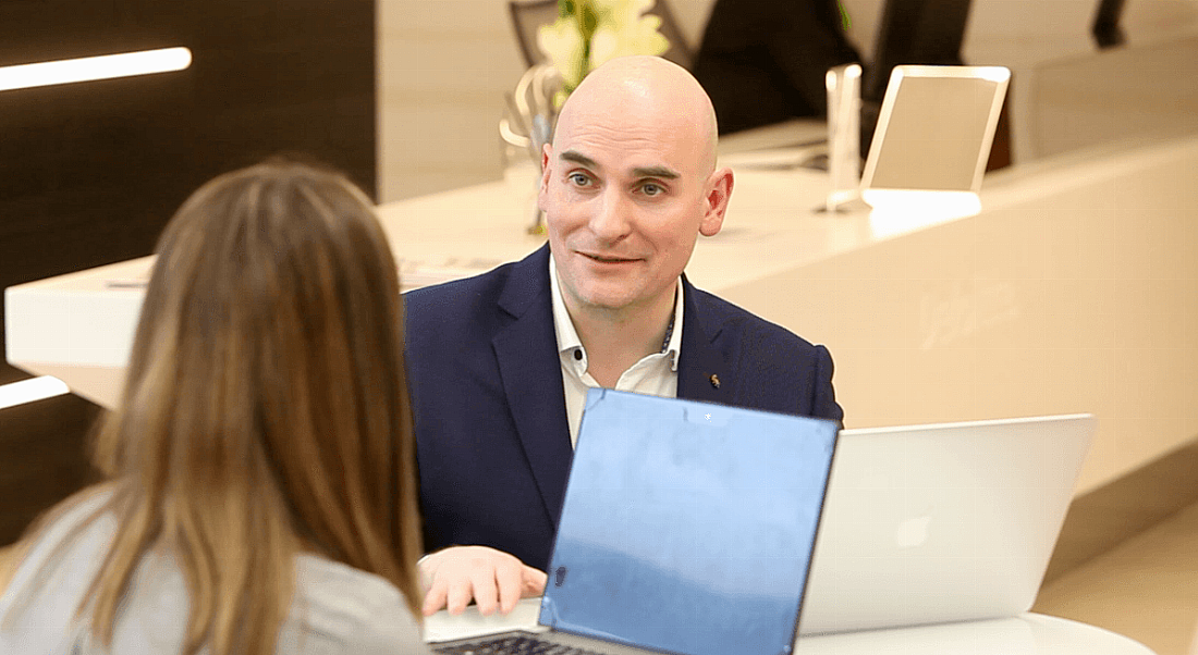 A bald man in a suit sitting with a laptop and talking to a brown-haired woman. They are representing agile working in Deloitte.