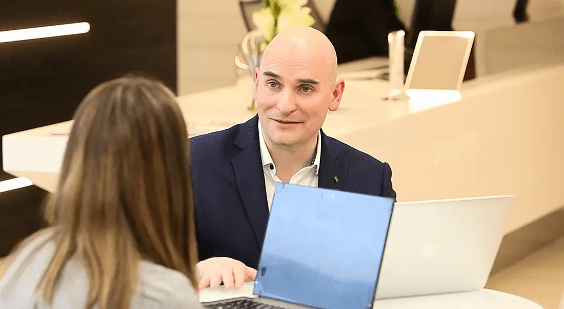 A bald man in a suit sitting with a laptop and talking to a brown-haired woman. They are representing agile working in Deloitte.