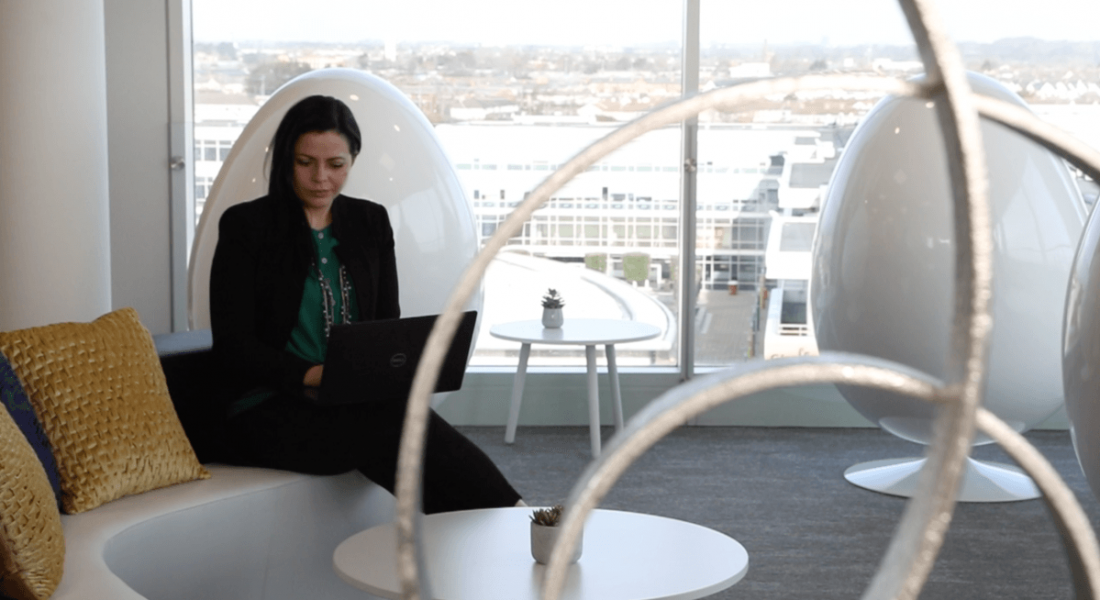 A woman in smart casual attire sitting on a couch working on a laptop in a well-lit office space.