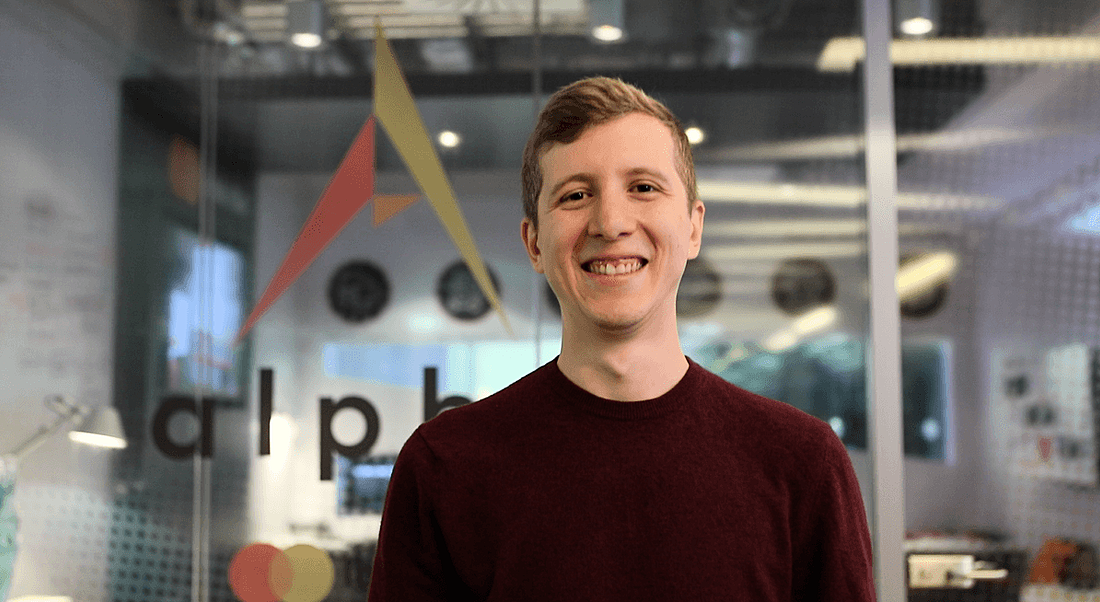 A young blonde man in a wine-coloured sweater gives the camera a toothy smile, head angled slightly downwards.