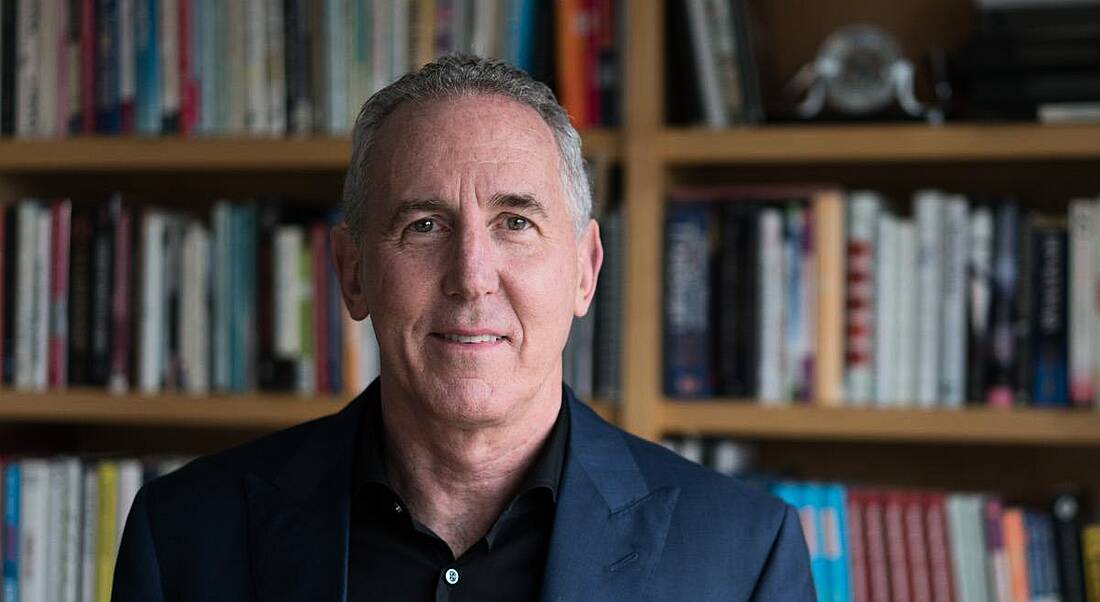 A man in a blue blazer and black shirt standing in front of a library full of books. He is from The Energy Project.