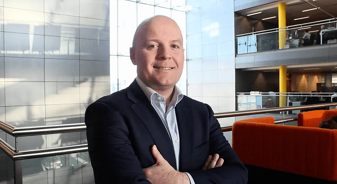 A bald man in a navy suit with his arms crossed smiling at the camera against a lofty office building backdrop.