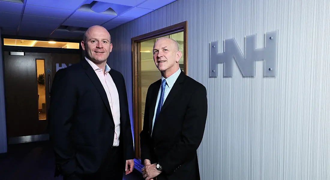 Two middle aged men in dark, crisply pressed suits standing next to each other smiling at the camera with HNH logo in background.