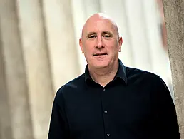 A man in a black sweater and white shirt leaning against a glass wall that is mirror reflecting his image.