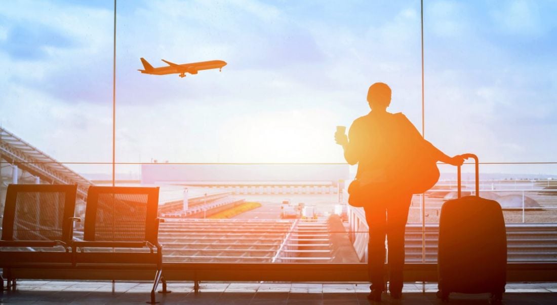 A man standing at an airport window with a suitcase looking at a plane representing someone with Stamp 3 visa.