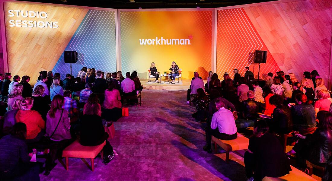 A shot of a small stage with the two women sitting in front of an audience. The Workhuman logo is behind them.