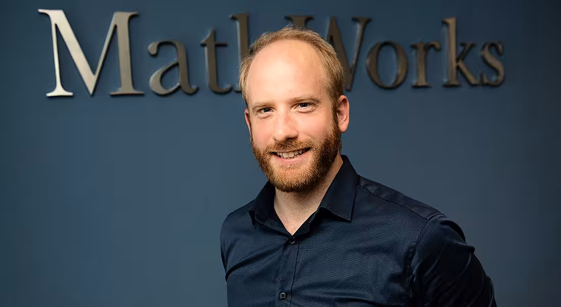 A professional man is smiling into the camera against a navy office wall with a sign reading 'MathWorks'.