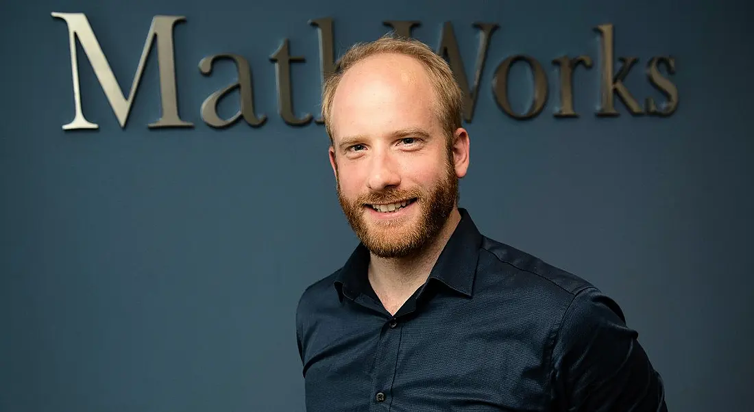 A professional man is smiling into the camera against a navy office wall with a sign reading 'MathWorks'.