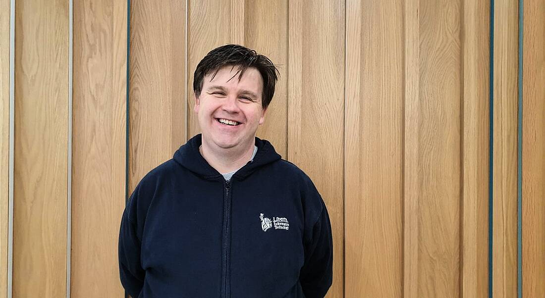 A man in a dark blue hoodie is standing against a wood-panelled wall and smiling into the camera.