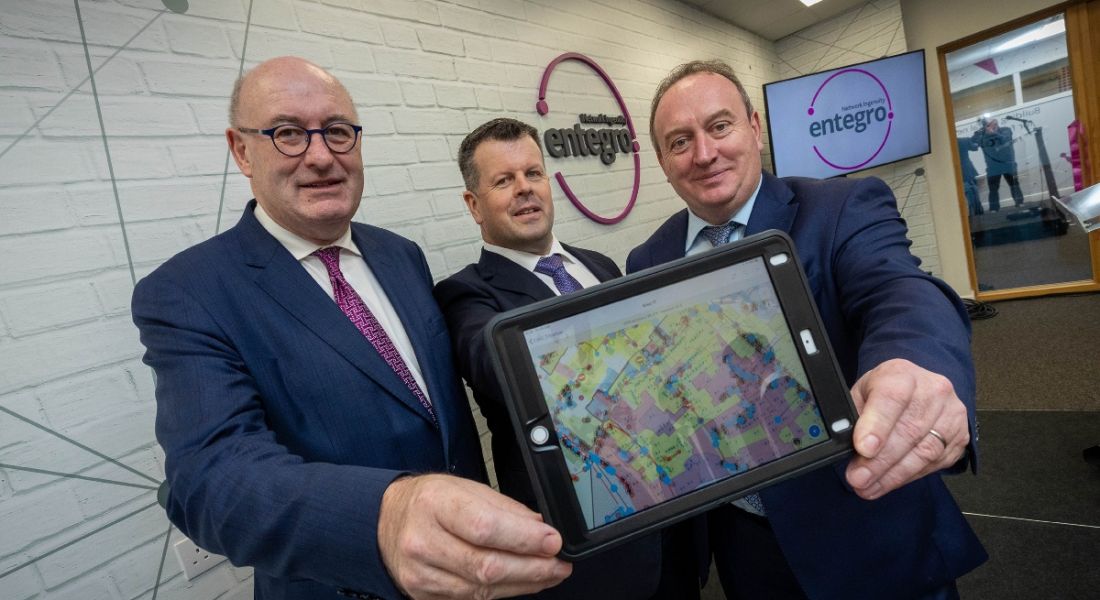 Three male business people in suits are looking at the camera in front of an Entegro sign on the wall, and holding a tablet with a digital display of mapped connections.