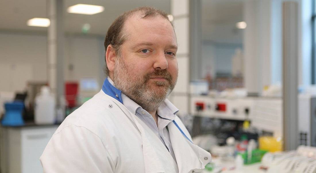 A man in a lab setting wearing a labcoat is looking into the camera.