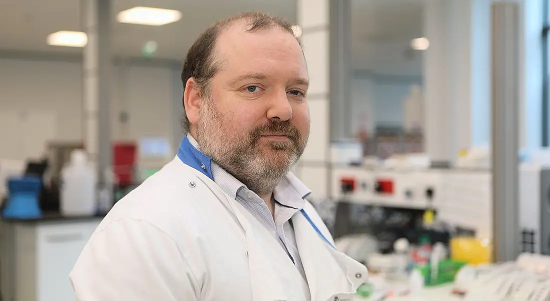 A man in a lab setting wearing a labcoat is looking into the camera.