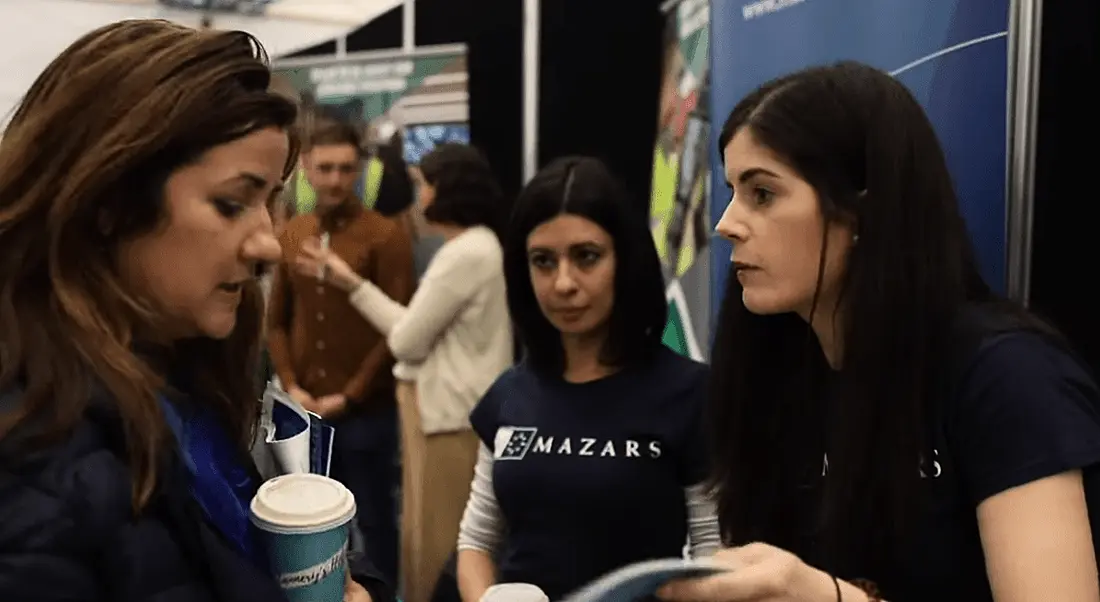 Three women interacting at a careers fair in an event hall.