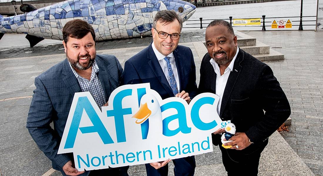 three businessmen holding an Aflac sign with a goose on it in an outdoors setting in Northern Ireland