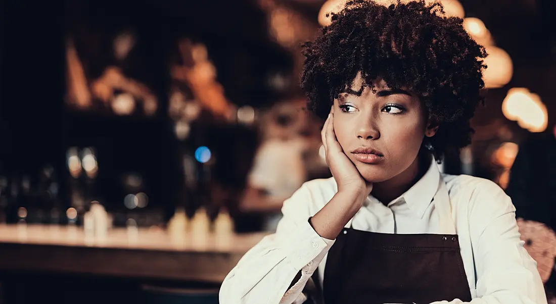 Young professional woman sitting at a table in a public café, looking demotivated with her cheek resting against her hand.