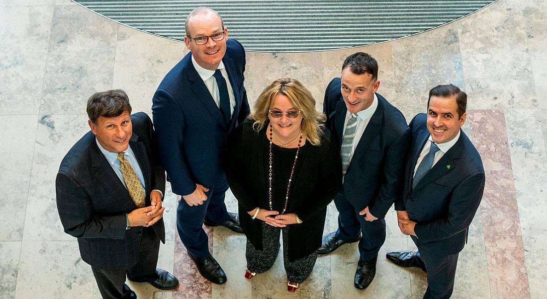 A group of four men in suits and one woman in business attire are standing together looking upwards at the camera and smiling.