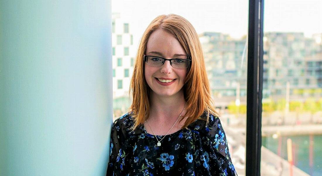 Photo of a young professional woman standing in front of a window looking onto Dublin's grand canal dock.