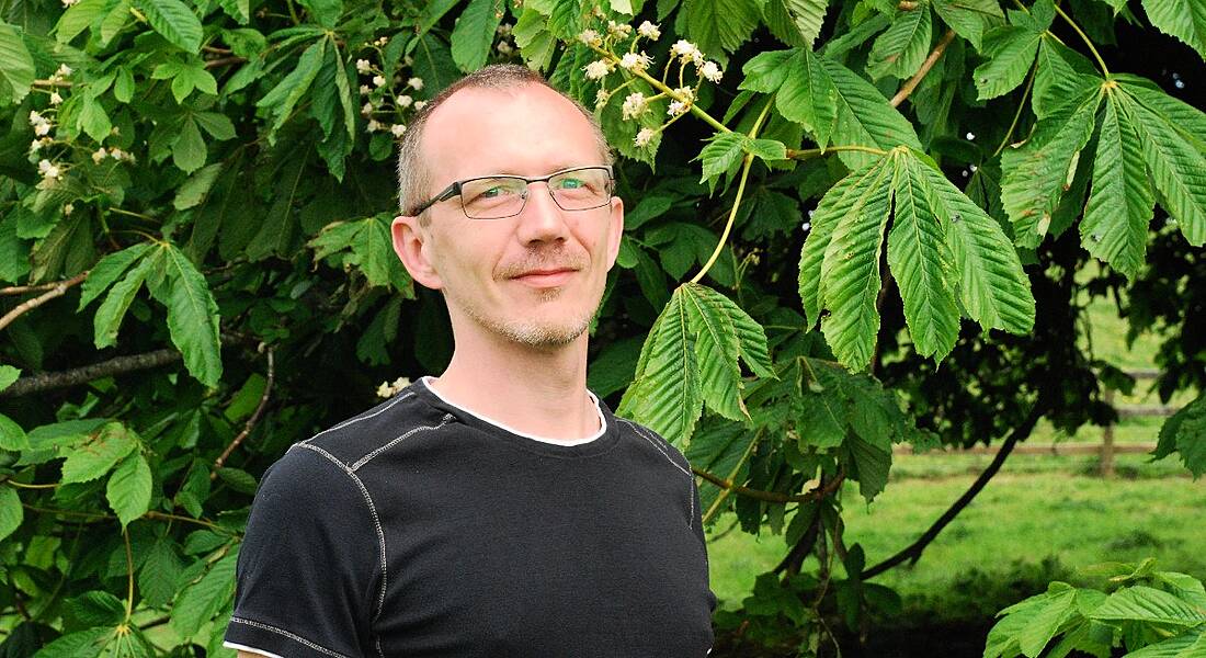 A man is standing against a backdrop of leaves outside, looking into the camera.