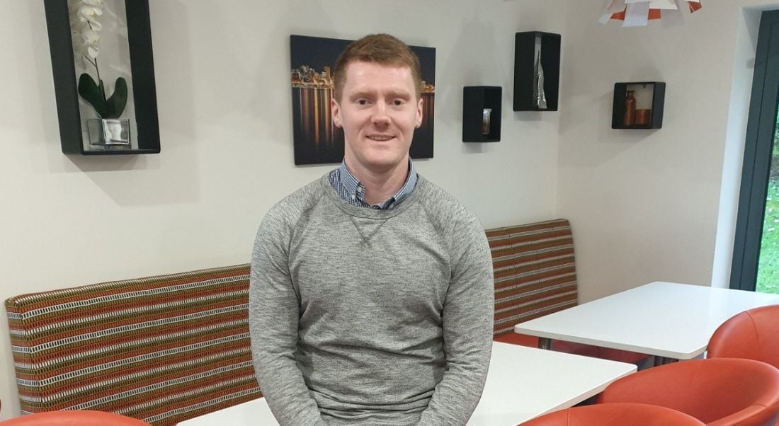 A professional man is smiling into the camera, leaning against a table in a communal working space at Storm Technology, Galway.