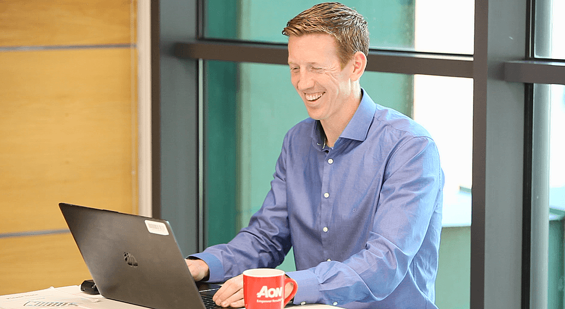 A man in business attire is sitting at a table using his laptop and smiling.