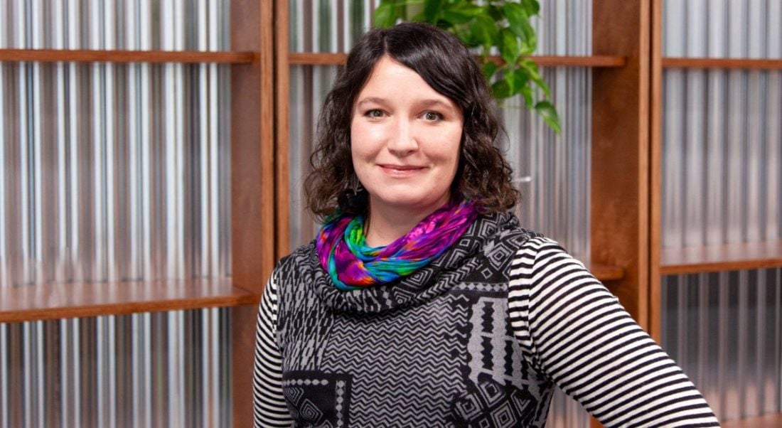 A woman with dark hair is smiling into the camera with one hand on her hip, against a contemporary office backdrop at Viasat.